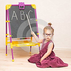 A little girl teacher in a dress and glasses sits on the floor near the blackboard and shows students the letters A B C