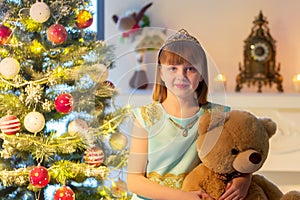 A little girl with a teddy bear is sitting on a sofa near a Christmas tree.