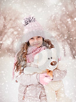 Little girl with a teddy bear stands on the street under the flying snow.