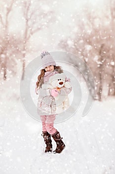 Little girl with a teddy bear stands on the street under the flying snow.