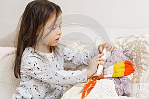 Little girl tie scarf around neck and measure temperature with electronic thermometer of sick teddy bear. Cure ill toy.