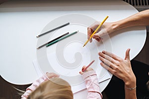 Little girl toddler with her mother drawing with colored pencils on table in children`s room at home, view from top