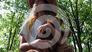 Little girl tourist in a hat with backpack and teddy bear is alone in the woods on sunny day. Child lost in the forest