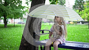 A little girl with a toy hare sits on a bench under an umbrella.