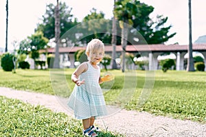 Little girl with a toy in her hand stands on the path on the lawn, half-turned