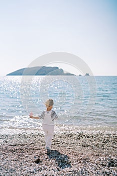 Little girl with a toy walks along a pebble beach towards the sea. Back view