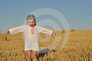 A little girl in a traditional chemise standing barefoot in a harvested field with ears in her hand
