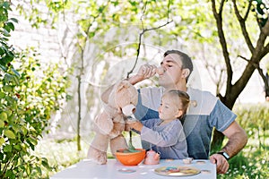 Little girl treats a teddy bear to tea from a toy cup while sitting next to her dad drinking from a toy cup