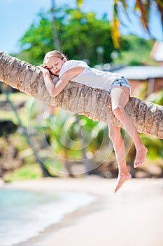 Adorable little girl at tropical beach sitting on palm tree during summer vacation