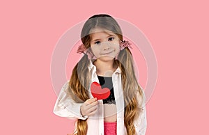 A little girl with two pigtails caught with two pink tops, who is pictured on a pink background holding a heart, is