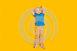 Little girl with two ponytails wears blue T-shirt and yellow pants. Child lying on bright yellow background. Top view
