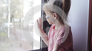 a little girl with a Ukrainian flag painted on her face is sad and looks out the window.