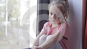 a little girl with a Ukrainian flag painted on her face is sad and looks out the window.