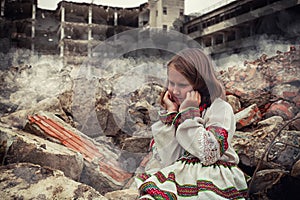 Little girl in Ukrainian folk costume sitting among ruins of house.