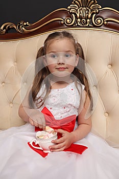 A little girl is waiting for the New Year and Christmas. A child in a white dress sits on a royal sofa with a cup of