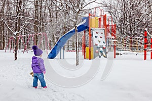 Little girl walking outside. Kid looking at the snowy children playground.