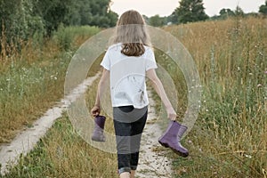 Little girl walking on rural road with rain boots in hands