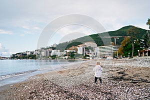 Little girl walks along the pebbly seashore against the backdrop of mountains and apartment buildings. Back view