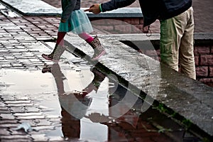 A little girl walks through a puddle in rubber baby boots. Dad holds his hand. Reflecting the child in the water