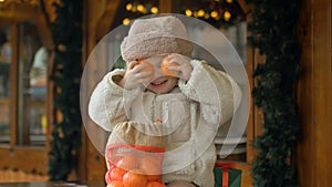 A little girl in warm clothes at the Christmas Fair. The girl jokingly covers her eyes with tangerines.