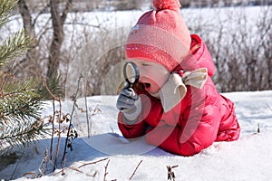 Little girl in warm winter clothes with a magnifying glass in her hand investigate  details of nature . Winter outdoor kids