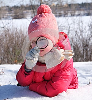 Little girl in warm winter clothes with a magnifying glass in her hand investigate  details of nature . Winter outdoor kids