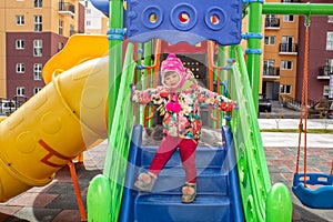The little girl, warmly dressed, in a hat and jacket plays on the playground with slides and swings in the courtyard of residentia