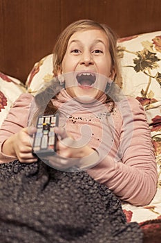 Little girl watching TV lying on bed with remote control in hand. vertical photo. toned