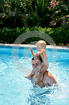 Little girl with a watermelon in her hand sits on the shoulders of her dad in the pool