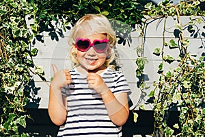 A little girl with wavy blonde hair in sunglasses holds her thumb with a like.