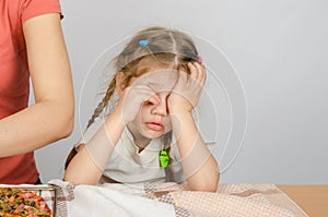 Little girl wearily rubbing his eyes at kitchen table while Mom cooks