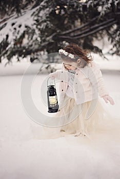 Little girl wearing a beige coat and long dress, looking at lamp with candle standing among the branches, covered with snow in the