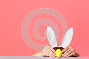 Little girl wearing bunny ears headband and playing with Easter egg at table against color background
