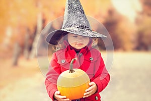 Little girl wearing Halloween witch hat and warm red coat, having fun in autumn day.