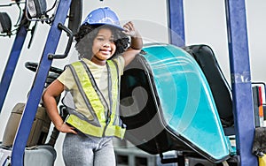 Little girl wearing hard hat, driving tractor in factory, smiling, playing alone as worker with happiness, doing activities on