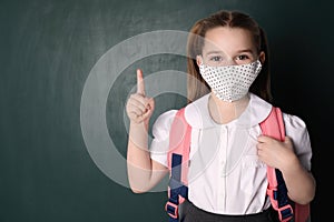 Little girl wearing mask and backpack near chalkboard, space for text. Child safety