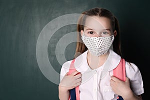 Little girl wearing mask and backpack near chalkboard, space for text. Child safety