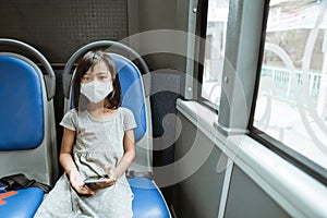 a little girl wearing a mask sits on a bench holding a cellphone on the bus