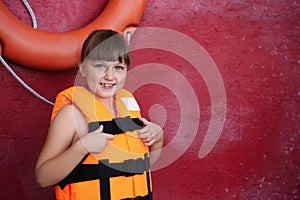 Little girl wearing orange life vest near red wall with safety ring. Space for text