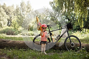 Little girl wearing pink protective helmet next to a bicycle with a back child bike seat. Safety while cycling with a
