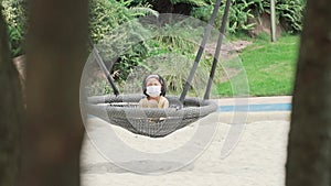 Little girl wearing protective face mask sitting on hanging chair in the garden
