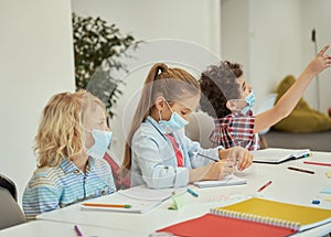 Little girl wearing protective mask looking at her notebook. Elementary school kids listening to teacher while sitting