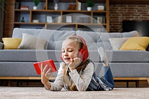 Little girl wearing red earphones lying on the carpet at home. Cute smiling child holding smartphone, watching cartoons