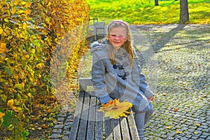 Little girl wearing retro coat and sitting on bench in park in autumn. Small girl is holding colorful autumn leaves. Autumn concep