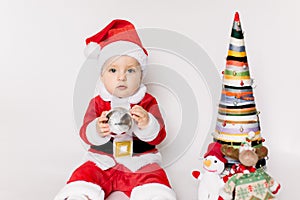 Little girl wearing santa claus red dress sitting on the floor holding small Christmas ball and smiles over white background,