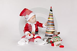 Little girl wearing santa claus red dress sitting on the floor holding small Christmas ball and smiles over white background,