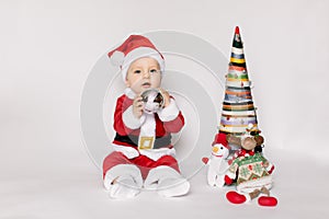 Little girl wearing santa claus red dress sitting on the floor holding small Christmas ball and smiles over white background,