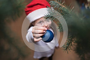 Little girl wearing Santa hat decorating Christmas tree with toys for Christmas in living room at home. Focus on the Christmas