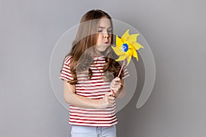 Little girl wearing striped T-shirt blowing at paper windmill, playing with pinwheel toy on stick.