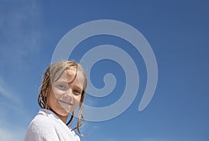 A little girl with wet blond hair, wrapped in a white towel, smiles against the blue sky. Summer vacation at sea. Happy Holidays